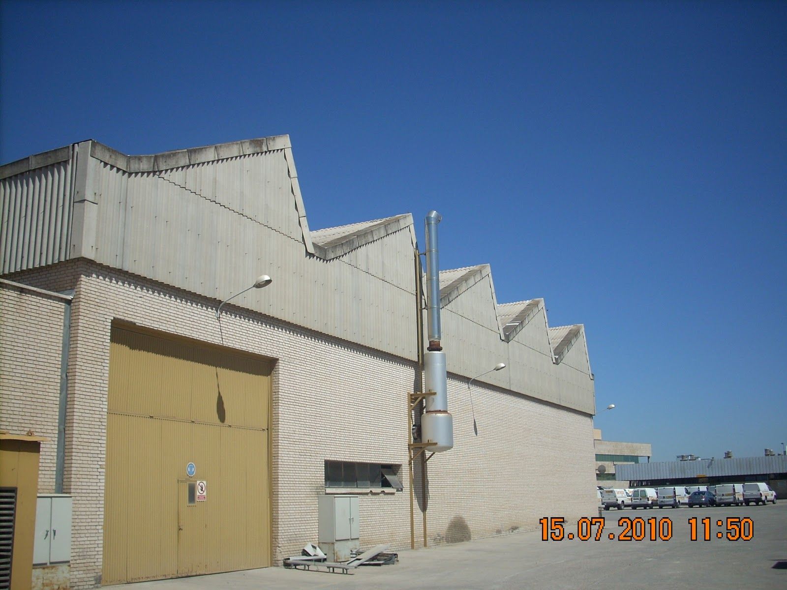 Exterior del almacén con techo de dientes de sierra, puerta grande y tubo de escape contra un cielo azul.