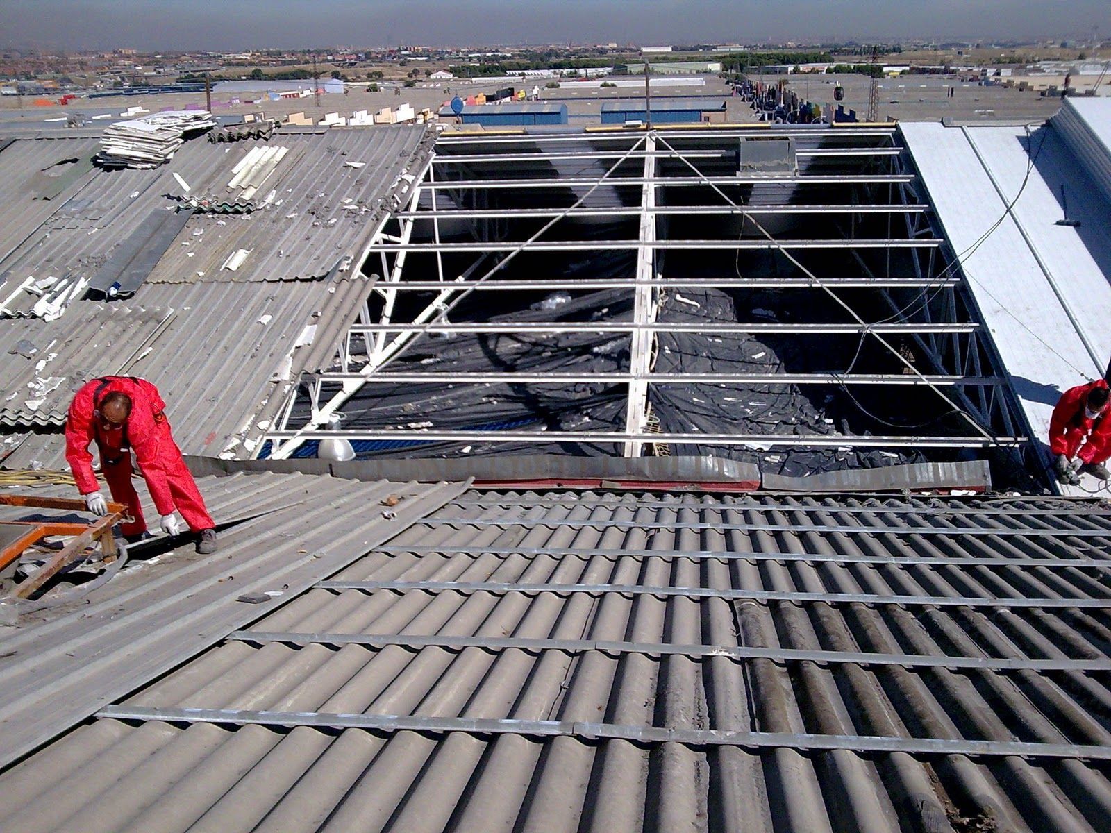 Dos trabajadores con overoles rojos reparan un techo de metal corrugado dañado, con una gran sección abierta.