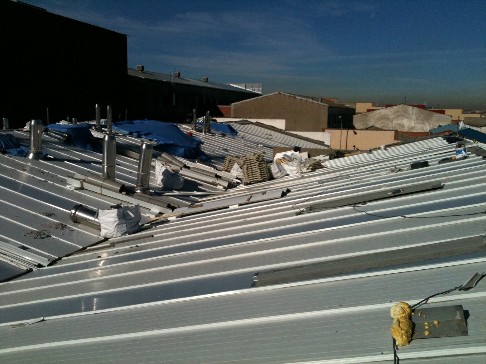 Techo metálico industrial dañado con escombros, respiraderos y un cielo parcialmente nublado.
