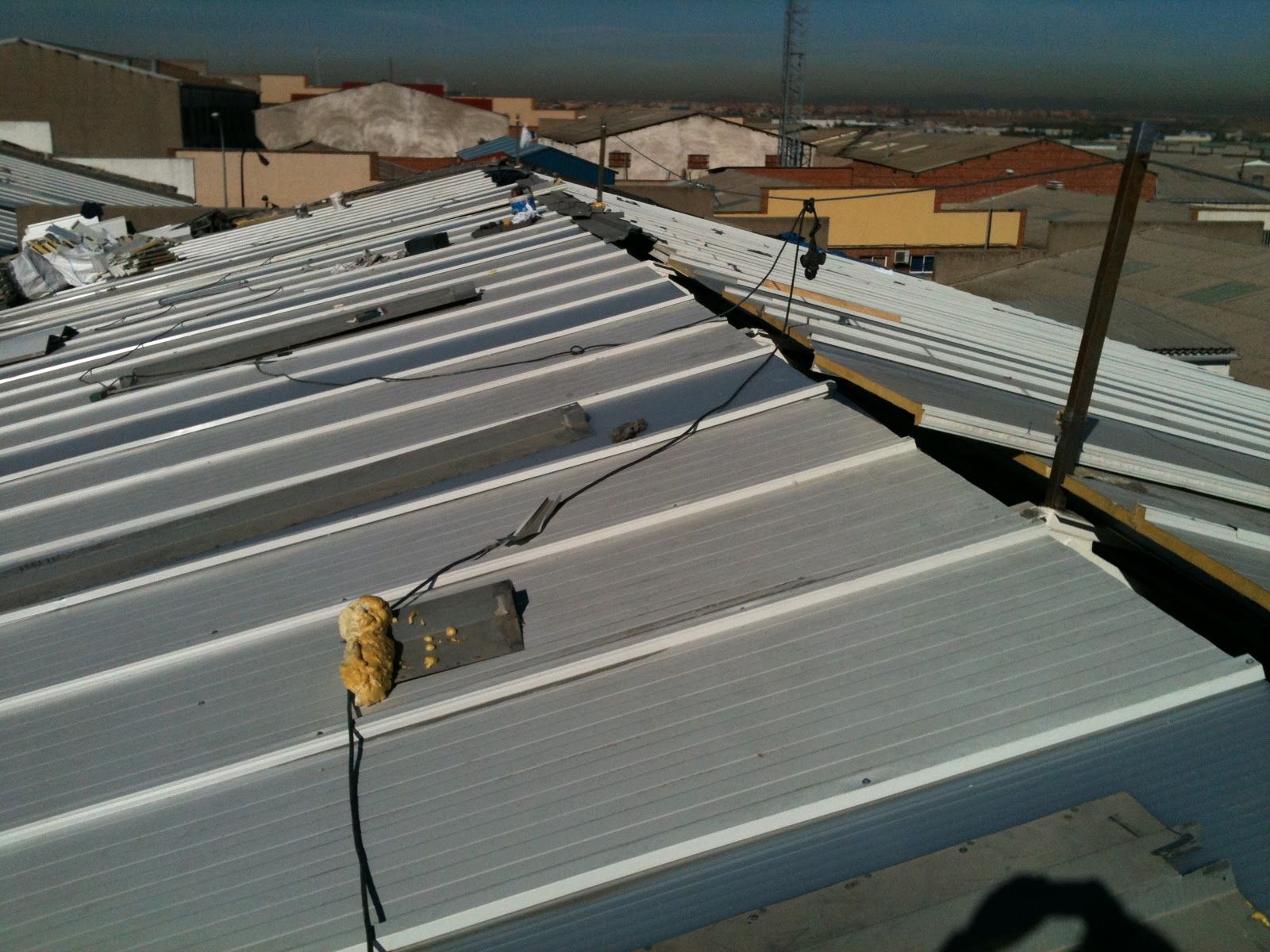 Techo metálico con equipo eléctrico y edificio al fondo. Exterior soleado.