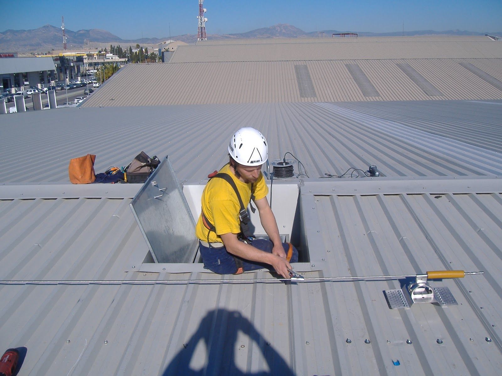Persona con un arnés en un techo de metal cerca de una escotilla abierta, trabajando con una barra de metal.