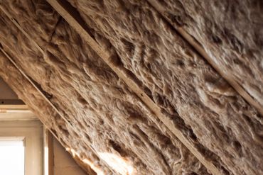Gros plan sur l'isolation d'un grenier incliné sous une charpente en bois, avec une petite fenêtre à gauche.