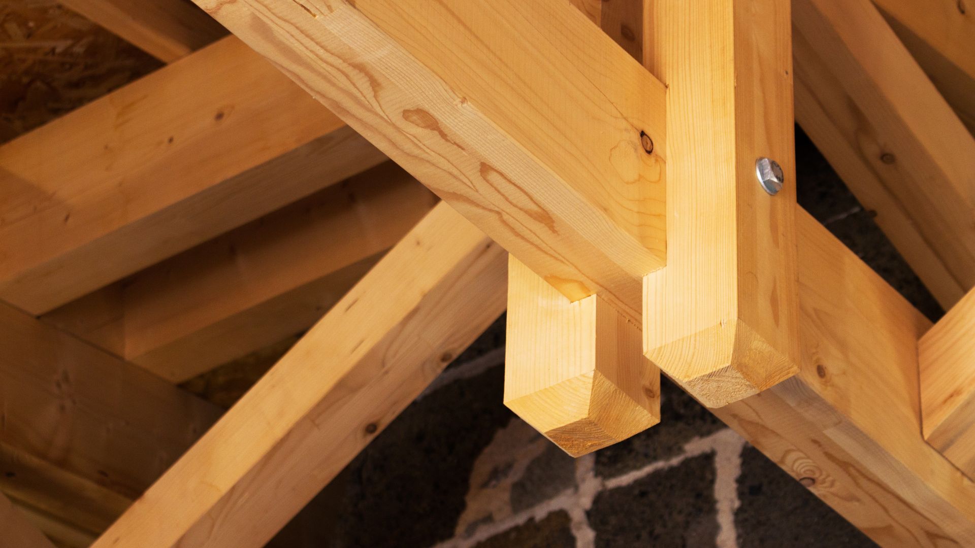 Gros plan sur des fermes de toit en bois apparentes, fixées par des boulons métalliques, contre un mur de pierre sombre.