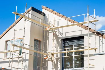 Extérieur d'une maison en construction, avec des échafaudages et des murs blancs sous un ciel bleu.