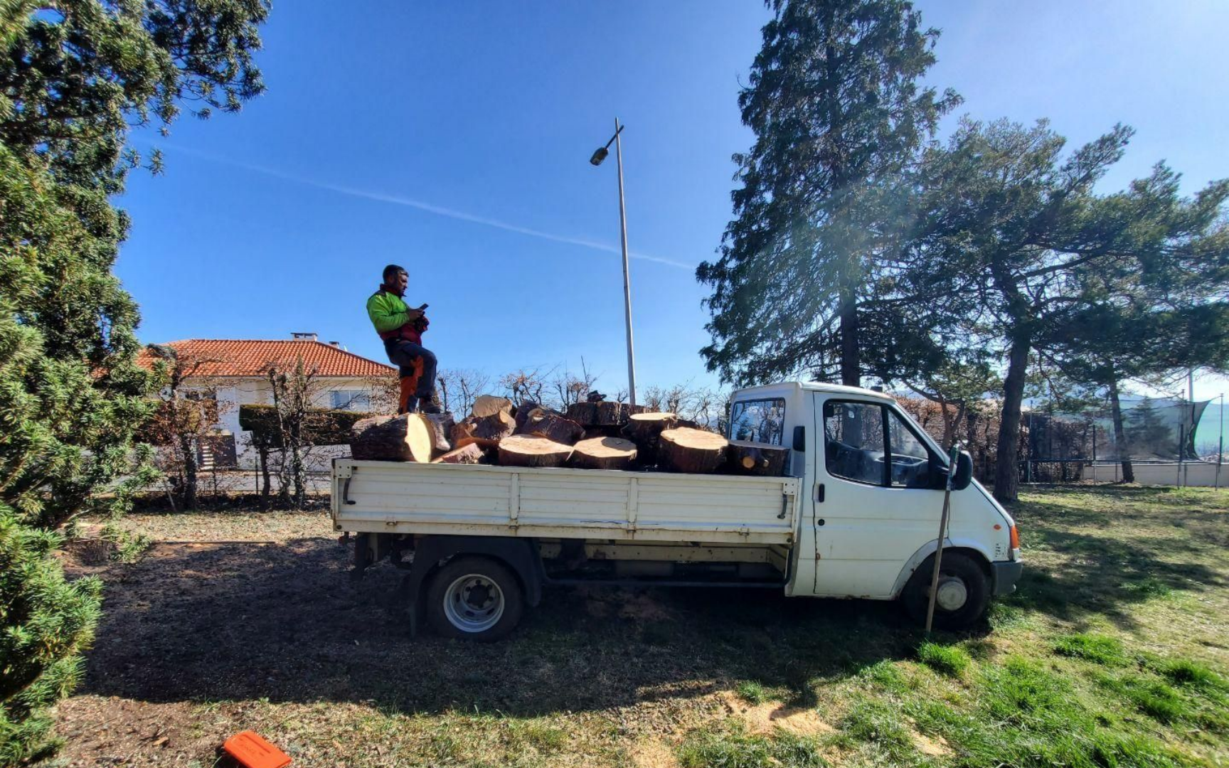 Un homme se tient debout à l'arrière d'un camion blanc chargé de grumes. Journée ensoleillée, ciel bleu, arbres et une maison en arrière-plan.