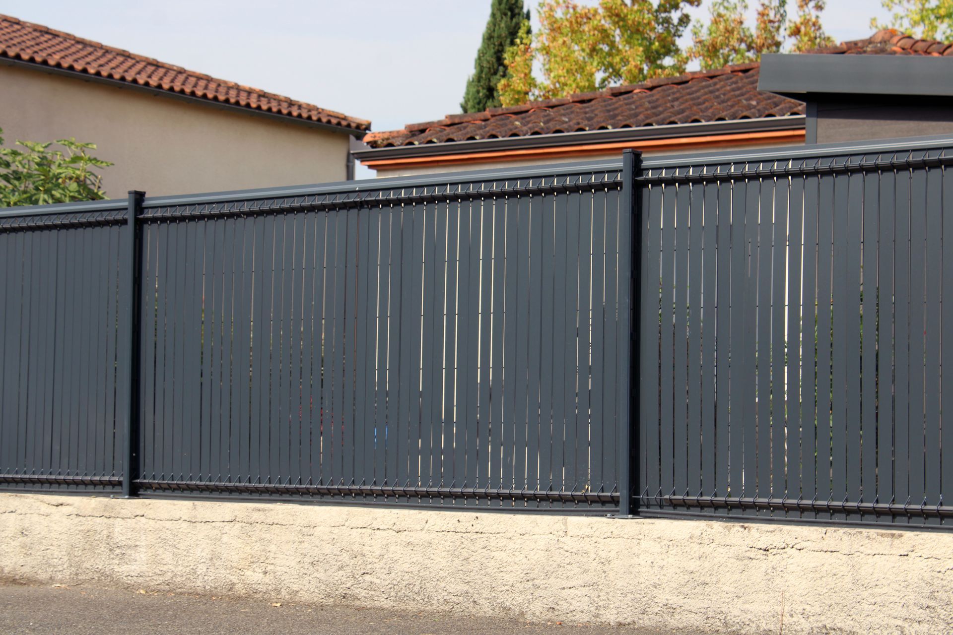 Clôture d'intimité gris foncé à fentes verticales, devant des maisons aux toits de tuiles rouges.