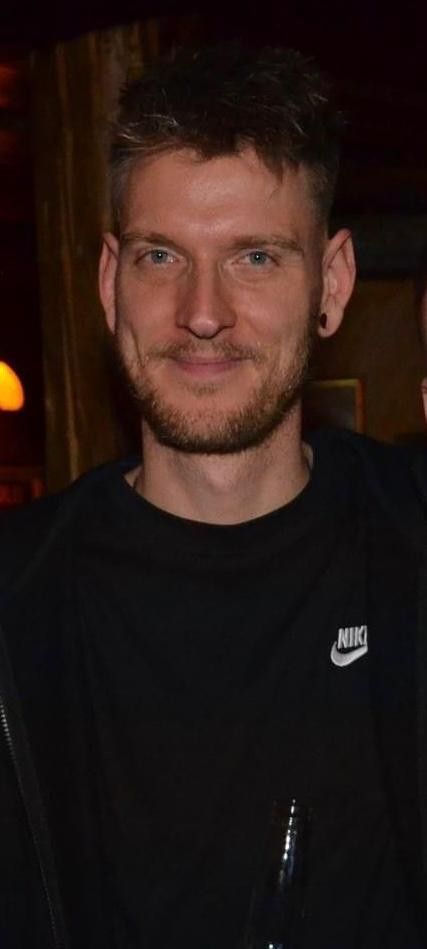 Ein Mann mit Bart trägt ein schwarzes Nike-Shirt und lächelt.