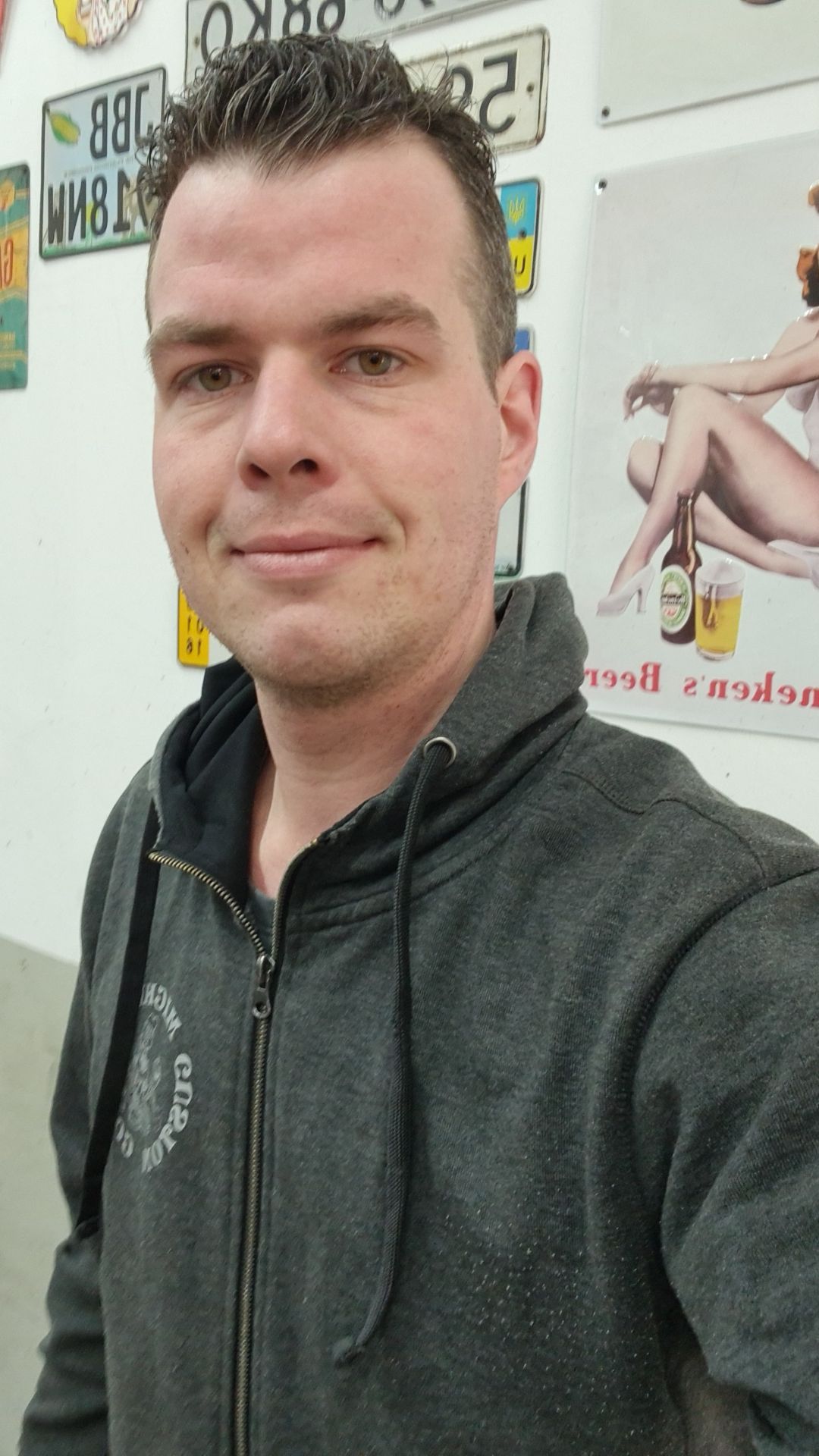 Ein Mann im Kapuzenpullover macht ein Selfie vor einer Wand mit Nummernschildern daran.