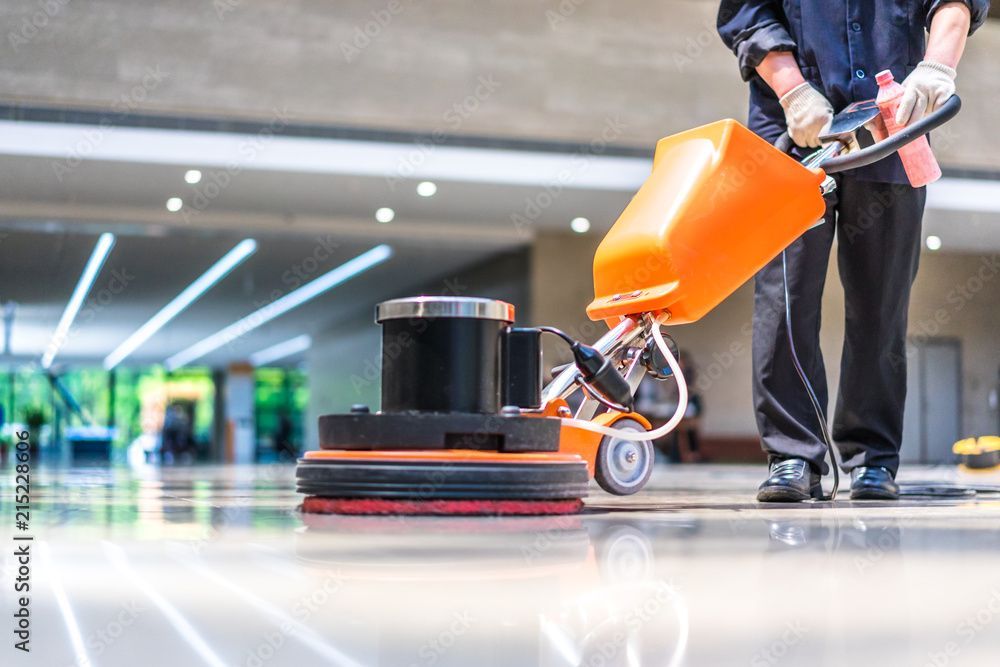 Eine Person benutzt eine orangefarbene Bodenschrubbmaschine in einer polierten, hell erleuchteten Lobby.