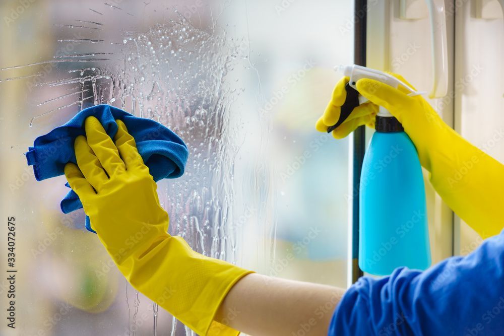 Mit gelben Handschuhen wird ein Fenster mit einem blauen Tuch und einer Sprühflasche gereinigt.