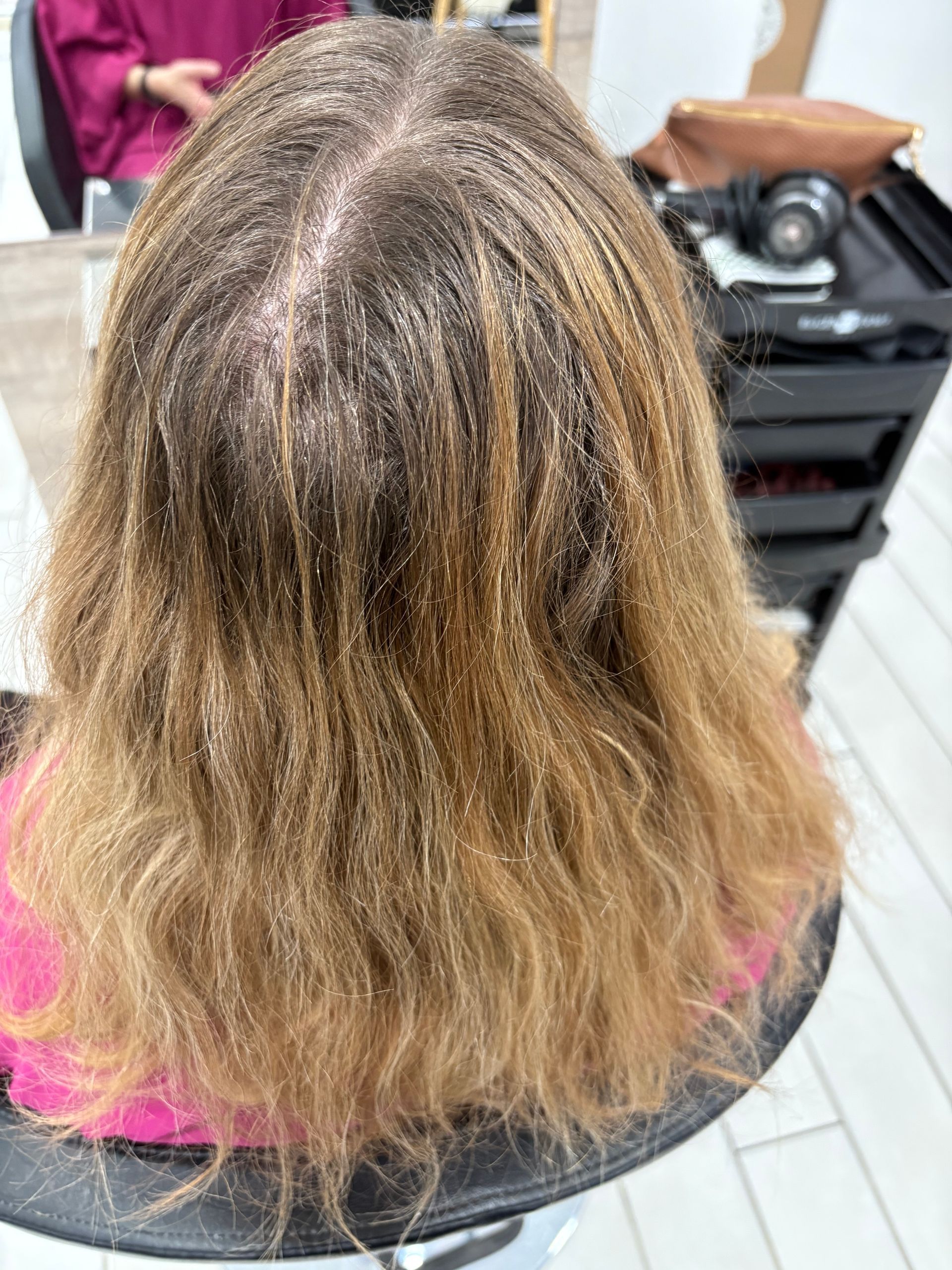 Cheveux d'une femme, raie au milieu, racines foncées et longueurs blond-brun, assise sur un fauteuil de salon.