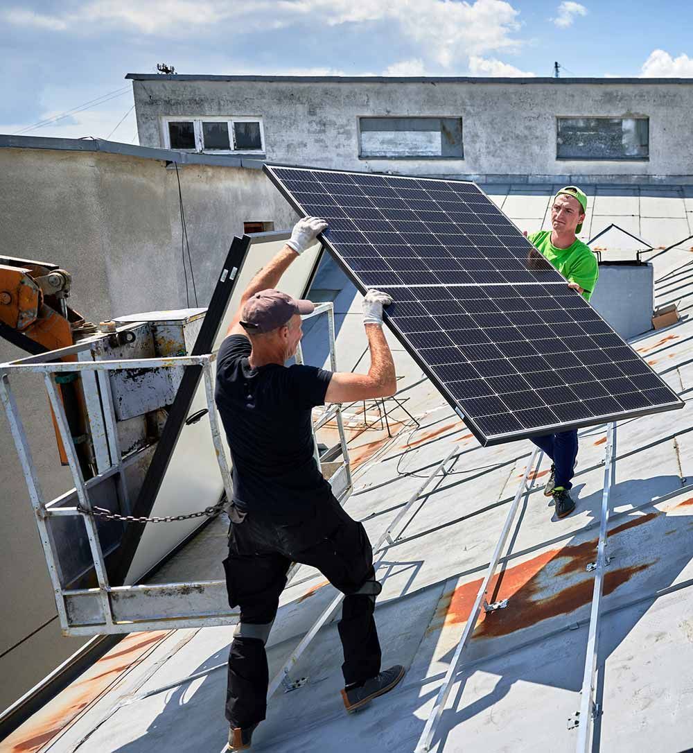Dos trabajadores instalan un panel solar en un tejado. Uno sostiene el panel y el otro ayuda.