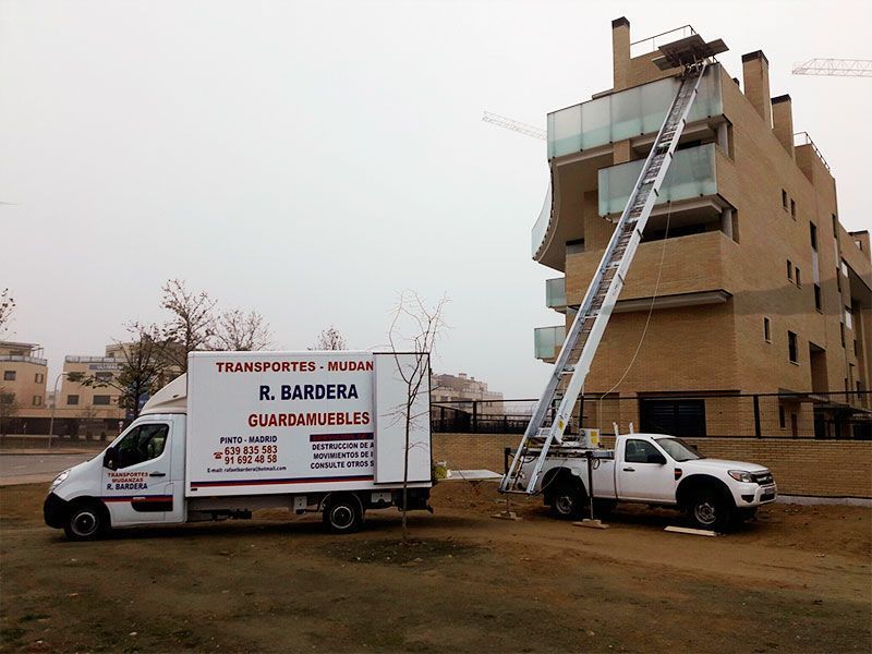 Camión de mudanzas blanco y camioneta con elevador exterior, cargando muebles en un edificio.
