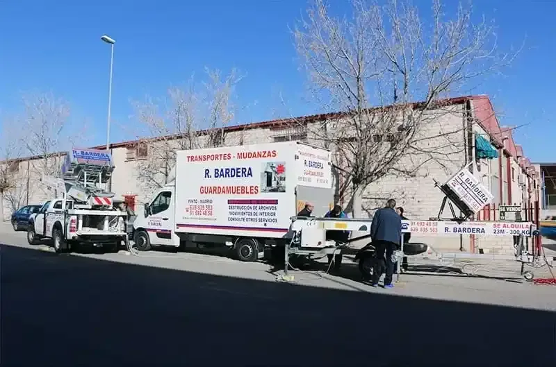 Camiones y equipo frente a un edificio. Trabajadores cargando y descargando. Cielo azul.