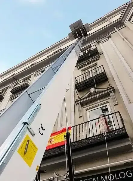 Un ascensor alto y en ángulo transporta mercancías a un edificio con balcones. Una bandera española cuelga debajo.