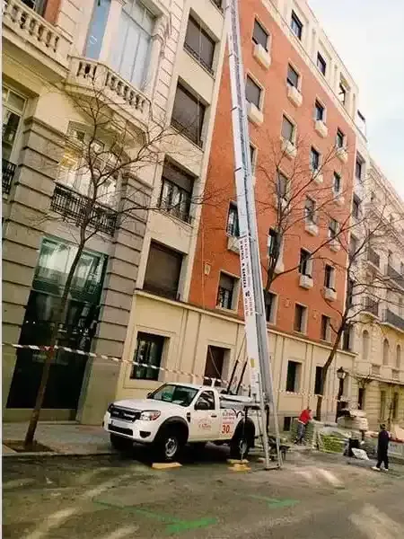 Camión blanco con elevador junto a un edificio. Hombres trabajando. Escena callejera con árboles y otros edificios.