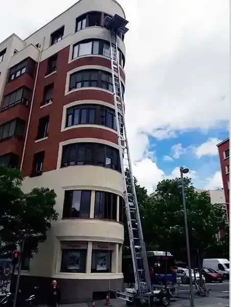 Una escalera alta que llega a un edificio, usada para mover objetos desde una ventana. Edificio marrón y blanco, cielo azul.