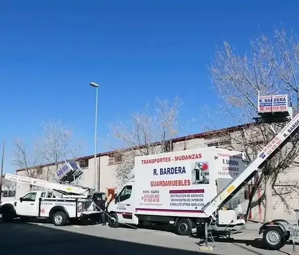 Dos camiones de mudanzas blancos con elevadores estacionados junto a un edificio bajo un cielo azul.