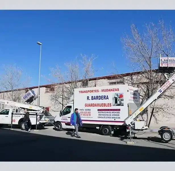 Camiones en movimiento con equipo elevador; una persona pasa por la calle.
