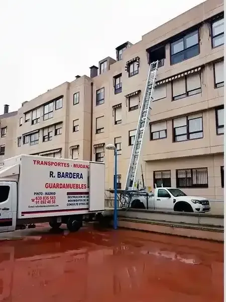 Camión de mudanzas con elevador extendido llegando a la ventana de un apartamento. Fachada del edificio visible.