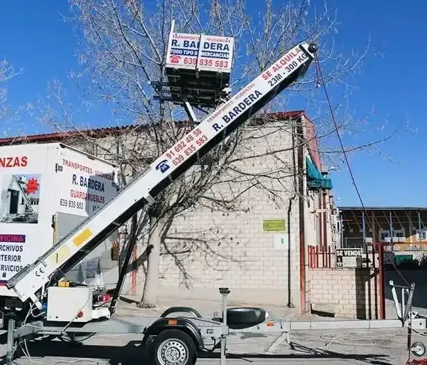 Un camión con plataforma elevadora extendida, cerca de un edificio y un árbol.