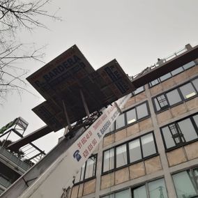 Un ascensor alto con plataformas, que se extiende hacia un edificio, probablemente para mover objetos. Cielo gris y nublado.