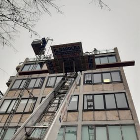 Un camión de mudanzas sube muebles a la azotea de un edificio de varios pisos. Una escalera se extiende hasta las ventanas.