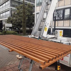 Listones de madera en una plataforma elevadora, exterior del edificio al fondo.