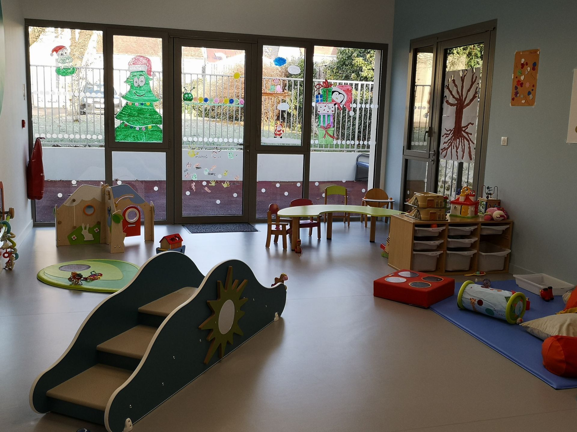 Salle d'éveil avec toboggan, petites tables, jouets et grandes fenêtres décorées.