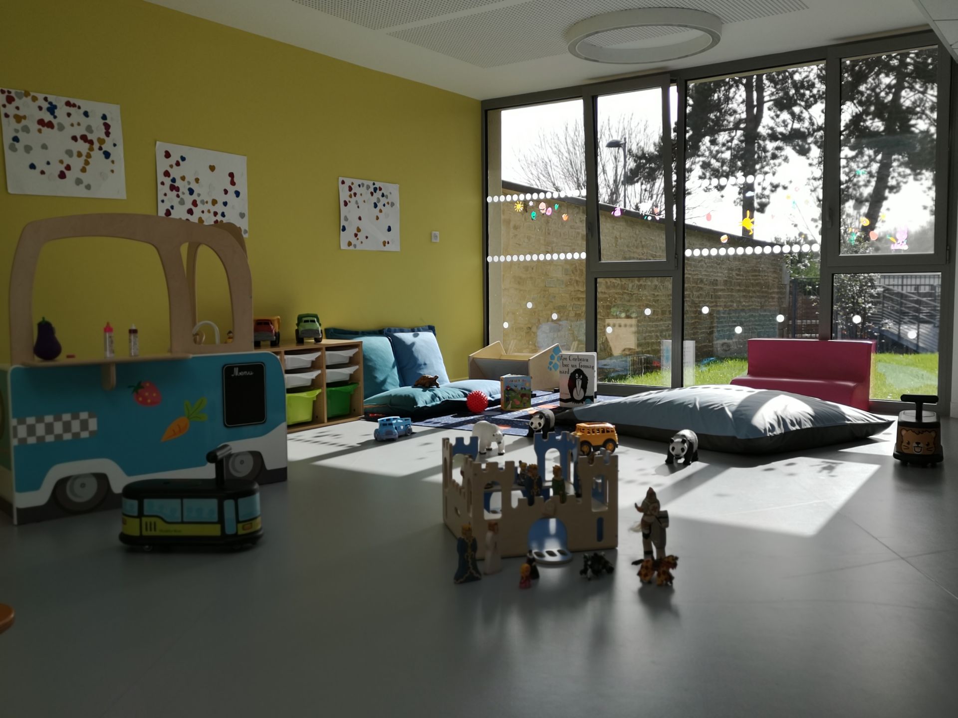 Salle de jeux avec des jouets éparpillés sur le sol, un canapé et une structure de jeu en forme de bus, grande fenêtre donnant sur l'extérieur.
