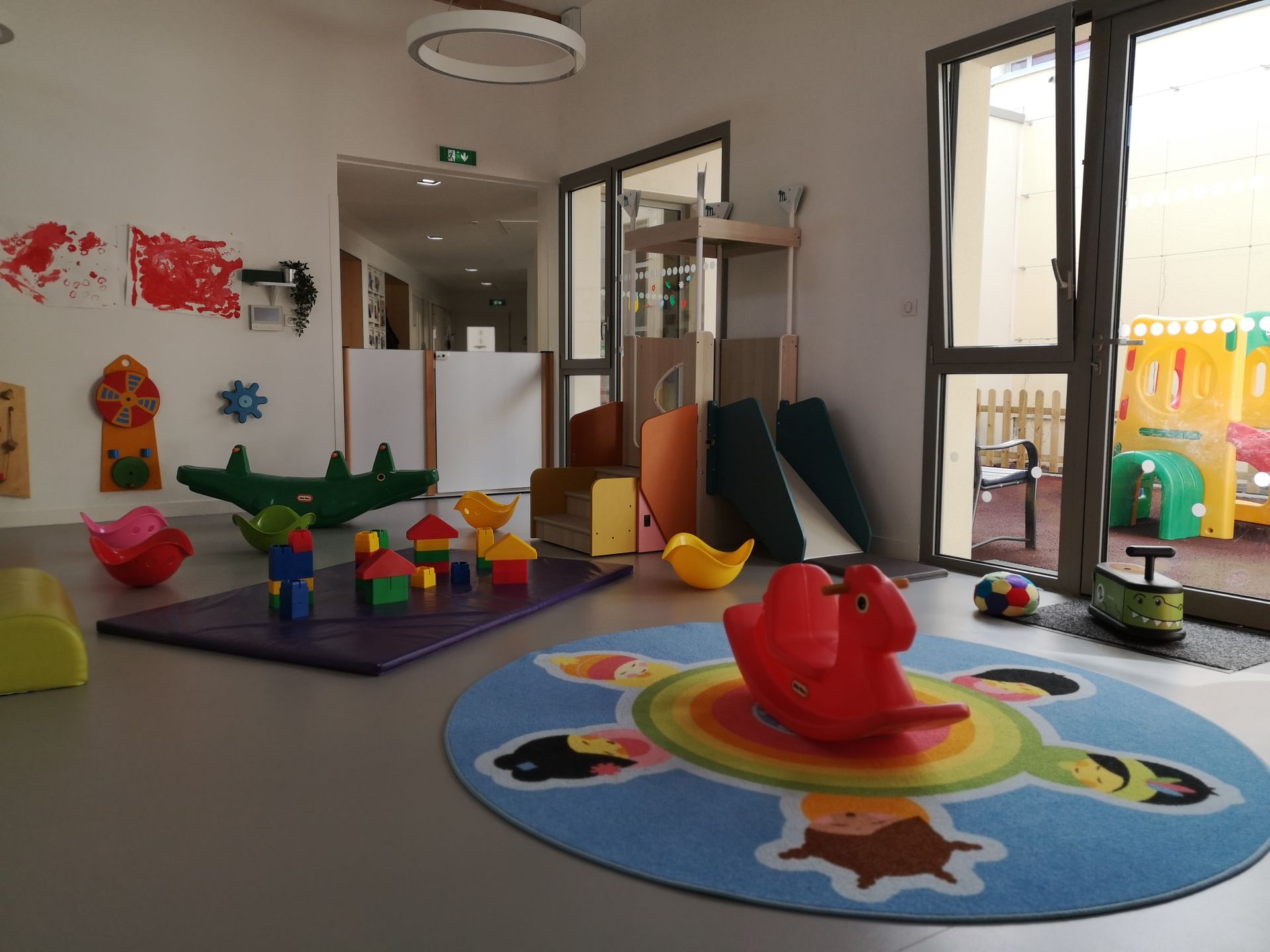 Intérieur de la salle de jeux avec des jouets, dont un cheval à bascule, et une aire de jeux extérieure visible.