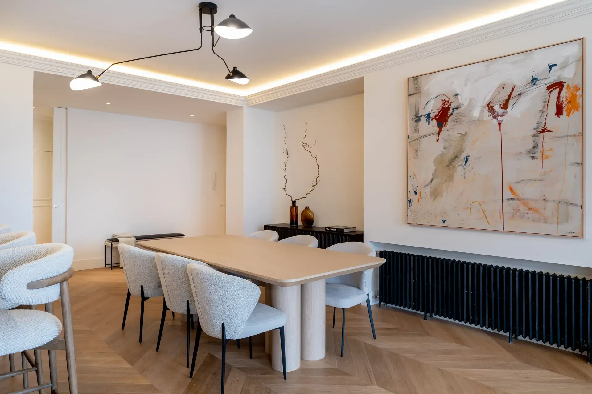 Comedor con una gran mesa beige, ocho sillas blancas, arte abstracto y lámpara moderna.