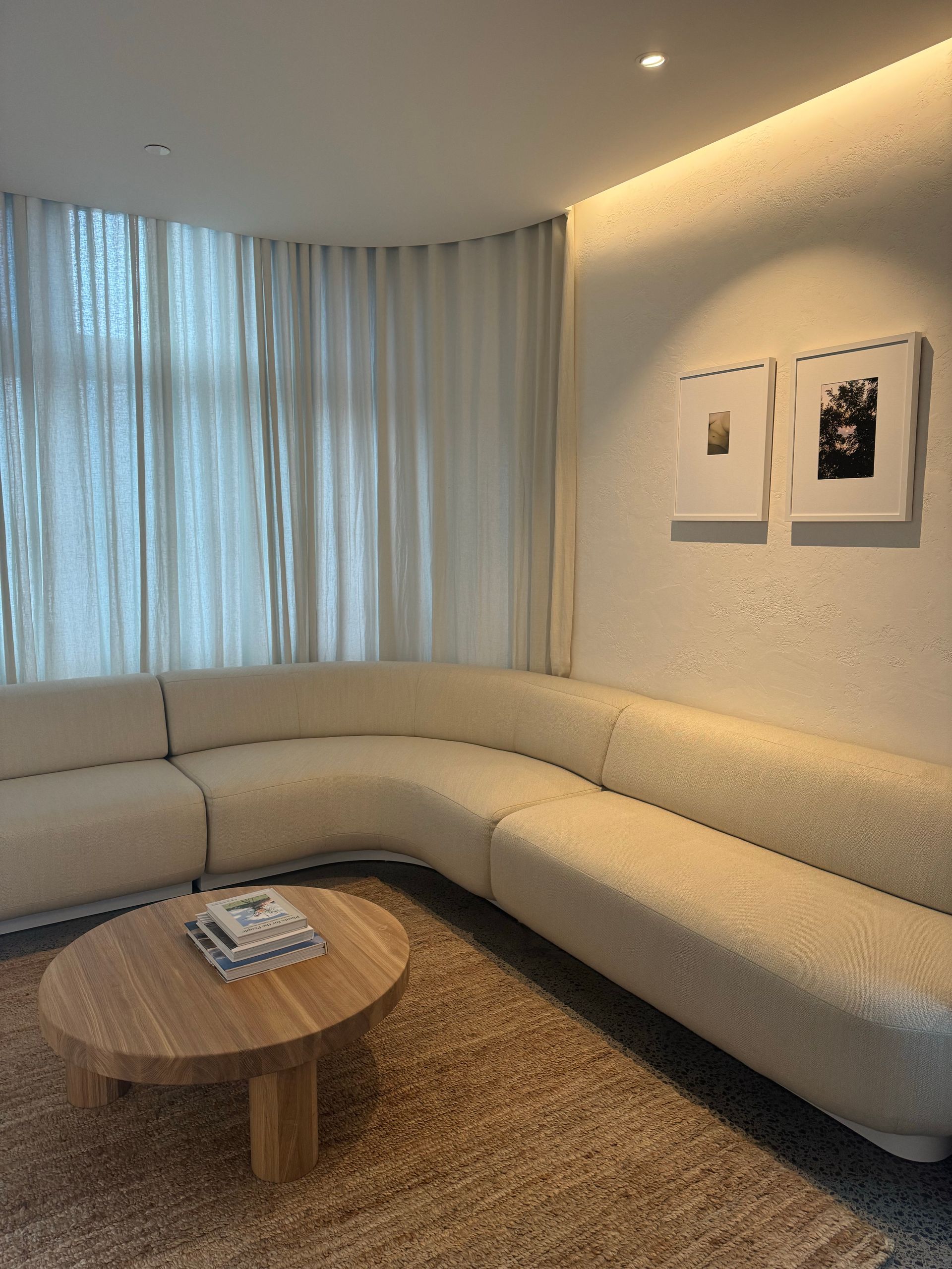 Sofá seccional color crema, mesa de centro redonda de madera y alfombra texturizada en una sala de estar luminosa. Cortinas blancas y cuadros enmarcados.