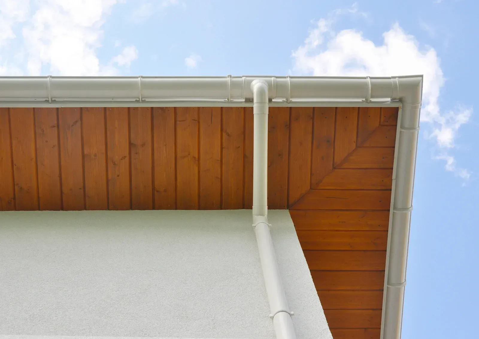 Roof edge with white gutter and downspout beside wooden soffit, against a blue sky.