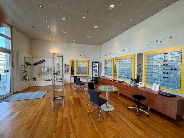 Innenansicht eines Optikergeschäfts. An den Wänden hängen Brillen. Zwei Sitzgruppen. Holzboden, weiße und gelbe Wände.