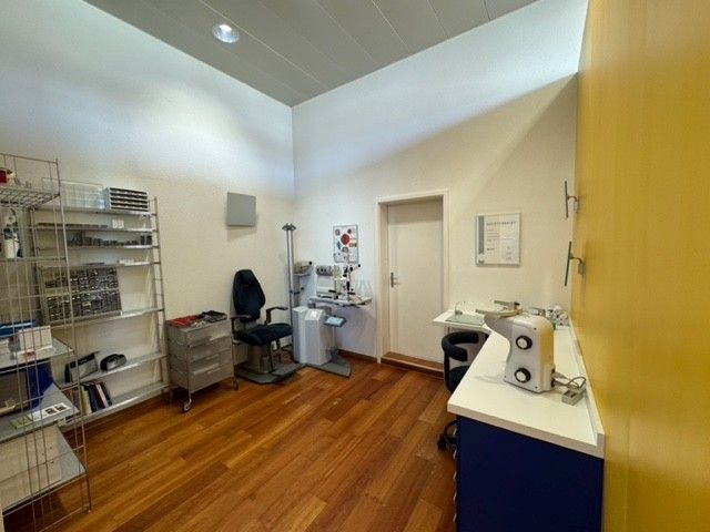 Ein Untersuchungsraum beim Optiker mit Geräten, einem Stuhl und einer Tür. Holzboden und gelb-weiße Wände.
