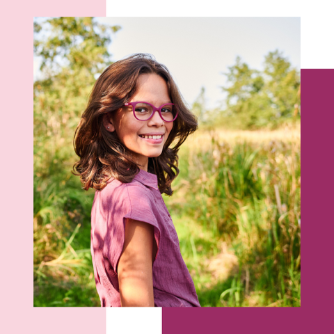 Mädchen mit rosa Brille und rosa Hemd lächelt auf einem Feld.