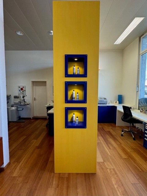 Gelbe Säule mit drei blau umrandeten Ausstellungsnischen, in denen jeweils weiße Flaschen stehen. Holzboden.
