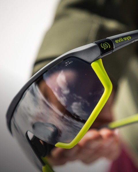 Nahaufnahme einer Sportsonnenbrille, graue Fassung mit neongelben Akzenten, die einen bewölkten Himmel reflektiert.