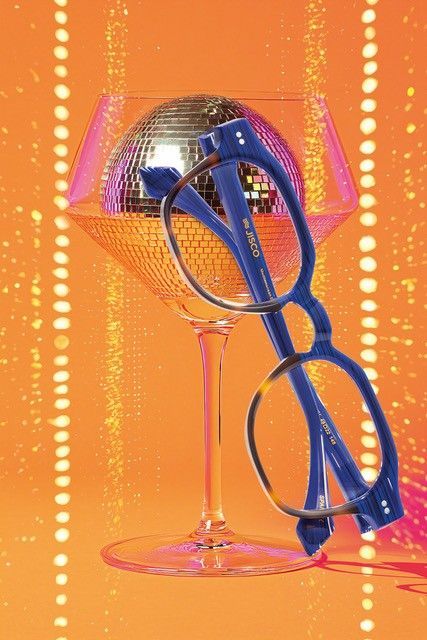 Eine blaue Brille liegt auf einem Champagnerglas mit einer Discokugel darin vor einem orangefarbenen, funkelnden Hintergrund.