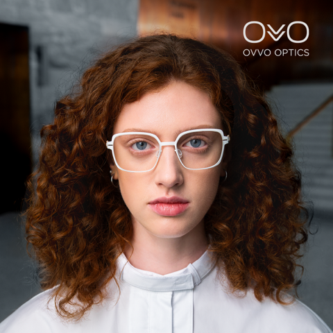 Frau mit lockigem, rotem Haar und weißer Brille blickt in die Kamera. OVVO Optics-Logo.