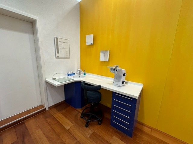 Augenuntersuchungsraum mit gelber Wand, weißem Schreibtisch und blauen Schränken. Enthält Untersuchungsgeräte.