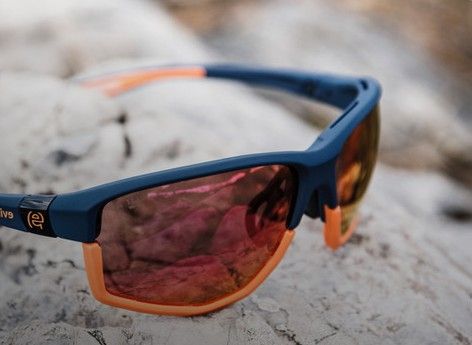 Blaue und orangefarbene Sonnenbrille auf strukturiertem, hellem Untergrund mit rosa getönten Gläsern.