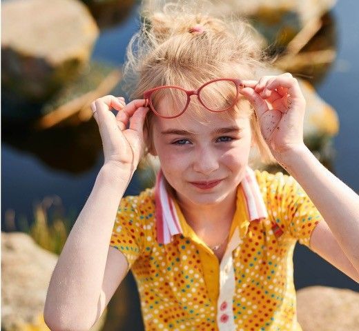 Mädchen mit heller Haut, trägt eine Brille, rückt diese mit den Händen zurecht, gelbes gepunktetes Hemd, draußen.