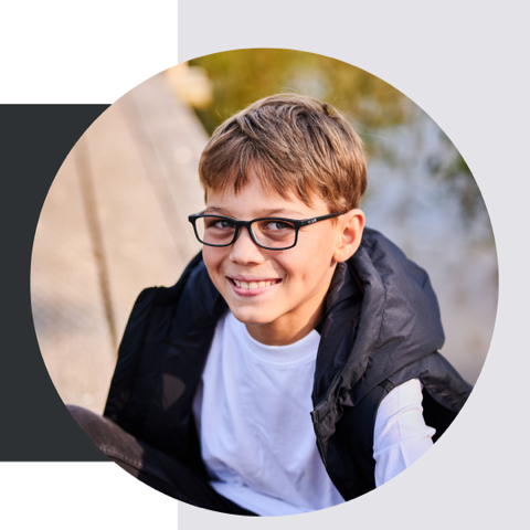 Ein Junge mit Brille lächelt; er trägt eine schwarze Weste über einem weißen Hemd. Draußen vor hellem Hintergrund.