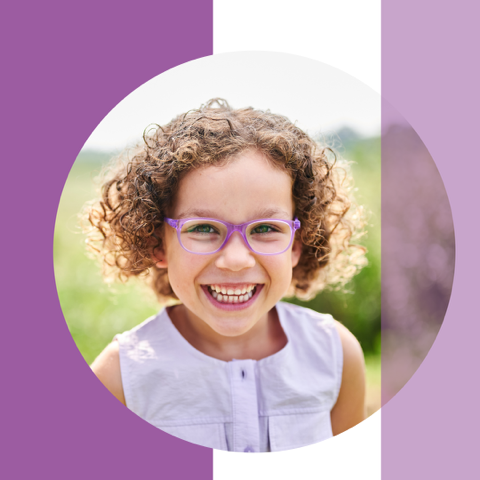 Lächelndes Kind mit lockigem Haar und lila Brille, draußen vor grünem Hintergrund.
