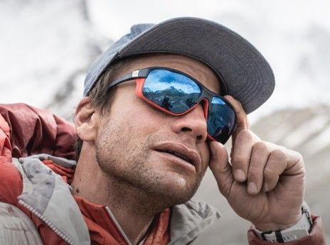 Mann mit Sonnenbrille und Kappe rückt seine Brille zurecht. Im Hintergrund schneebedeckte Berge.