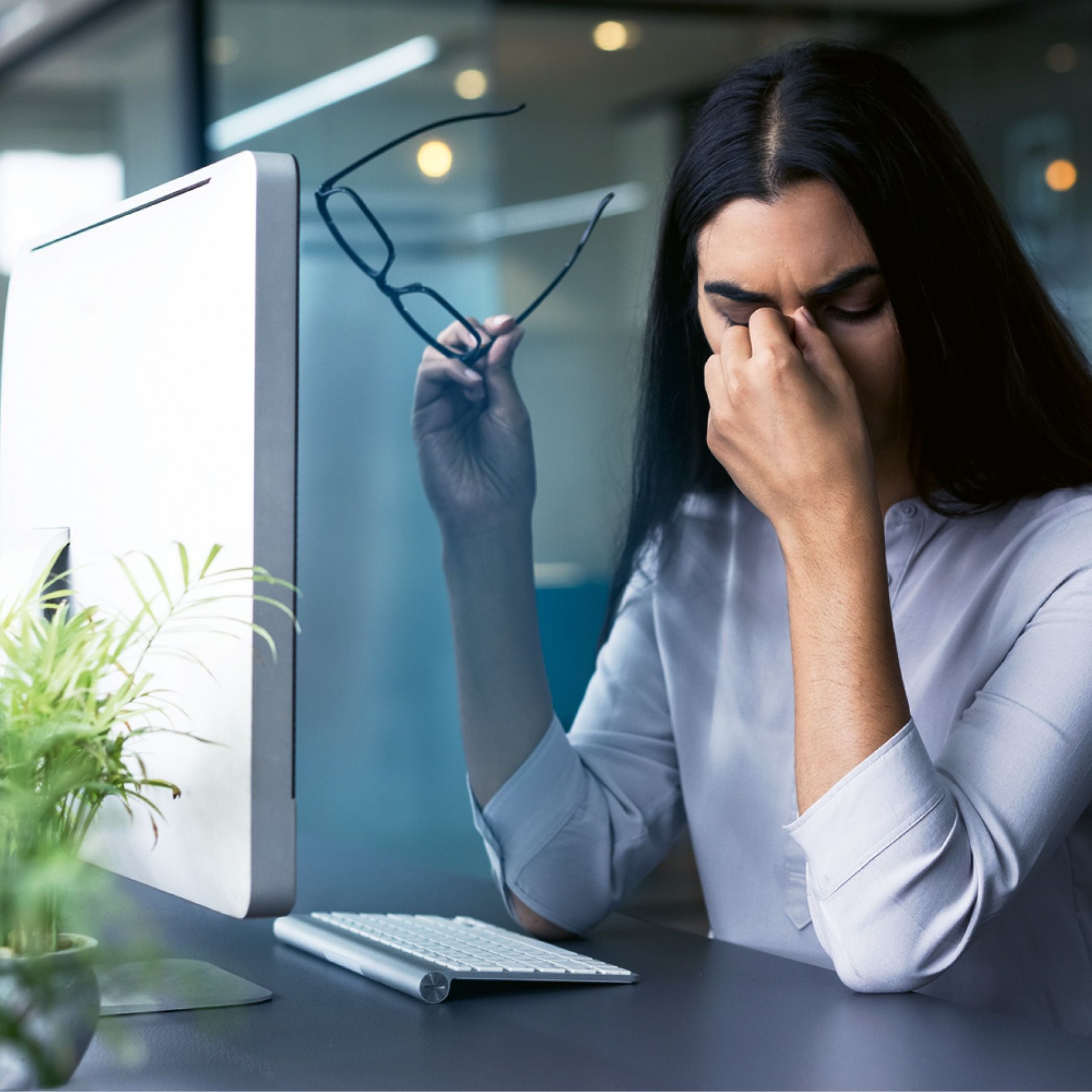 Frau am Schreibtisch, hält eine Brille, reibt sich die Augen, wirkt gestresst in der Nähe eines Computers.