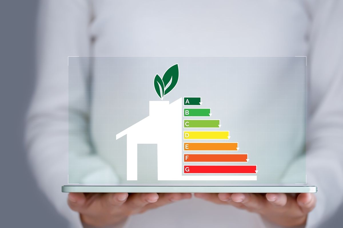 Persona sosteniendo una tableta con un ícono de casa, un cuadro de eficiencia energética y hojas verdes.
