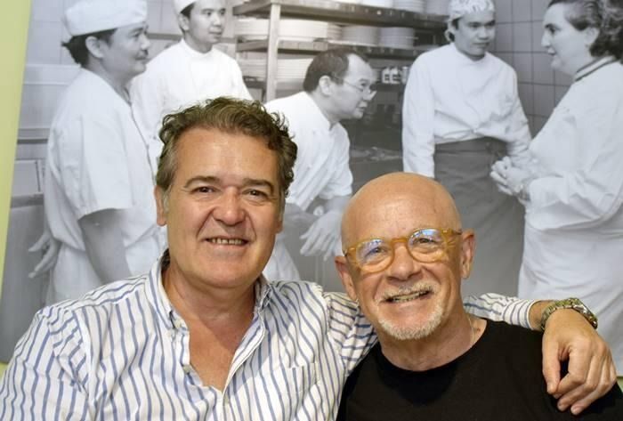 Dos hombres posan para una foto con chefs al fondo.